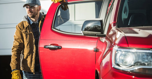 A man wearing work gloves opens the door of a red pickup truck parked beside a white garage in daylight.
