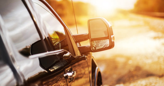 The side-view mirror of a black pickup truck reflects the sun setting over a bright roadside landscape.