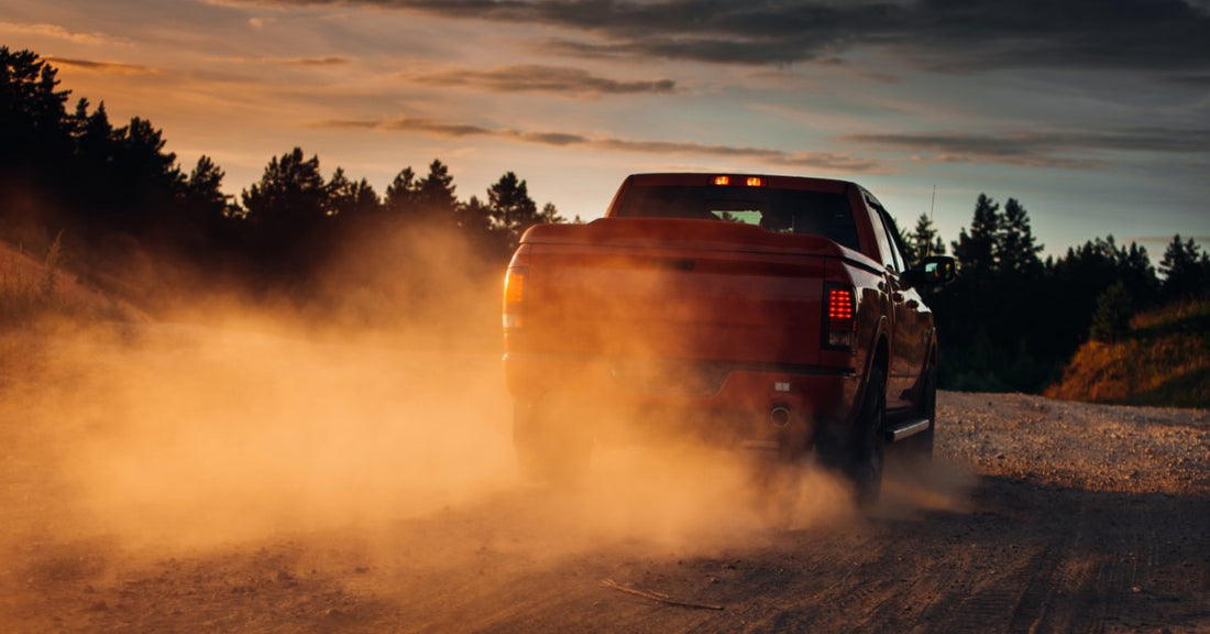 A red pickup creates a cloud of dust behind it as it drives down a gravel road beneath a dramatic sunset sky.