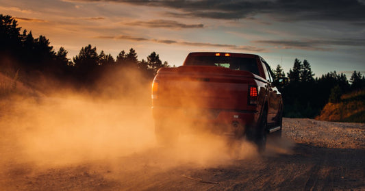 A red pickup creates a cloud of dust behind it as it drives down a gravel road beneath a dramatic sunset sky.