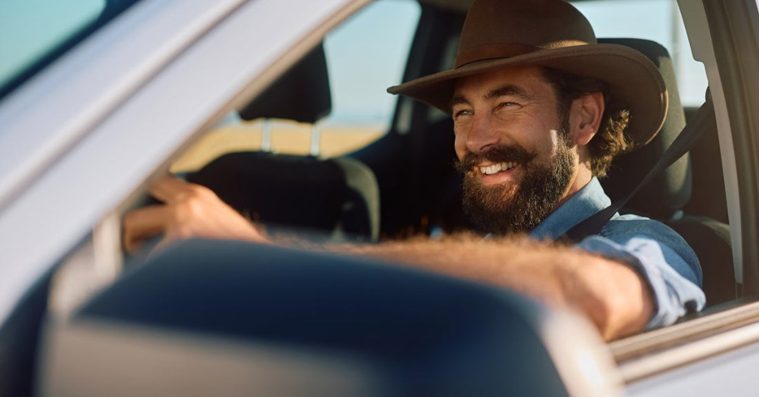 A smiling bearded man in a cowboy hat drives a white pickup truck with sunlight streaming through the window.