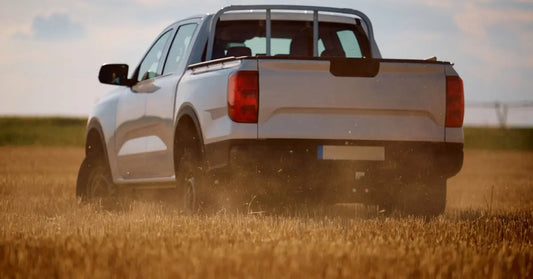 A white pickup truck drives through a golden field at sunset, kicking up dust behind its rear tires.