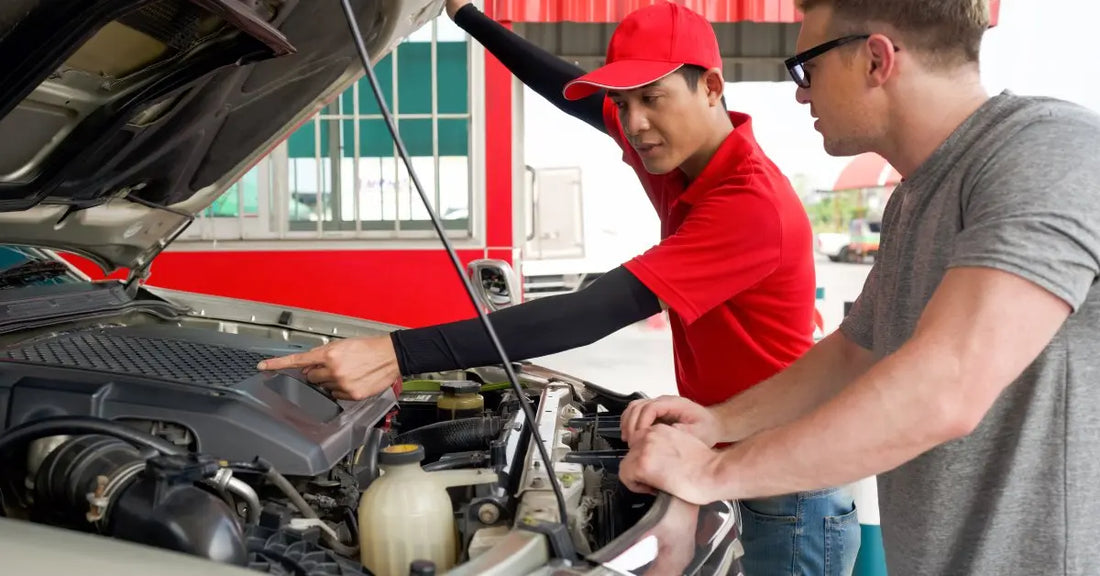 A mechanic and his customer standing next to the exposed engine bay of a pickup truck. The mechanic points to a part.