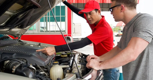 A mechanic and his customer standing next to the exposed engine bay of a pickup truck. The mechanic points to a part.
