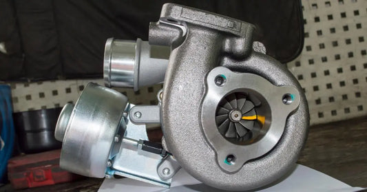 A silver turbocharger sits on a workbench, showing the turbine wheel through a round front inlet opening.
