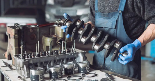 A mechanic in blue gloves holds a crankshaft over a workbench lined with pistons, rings, and an engine block.