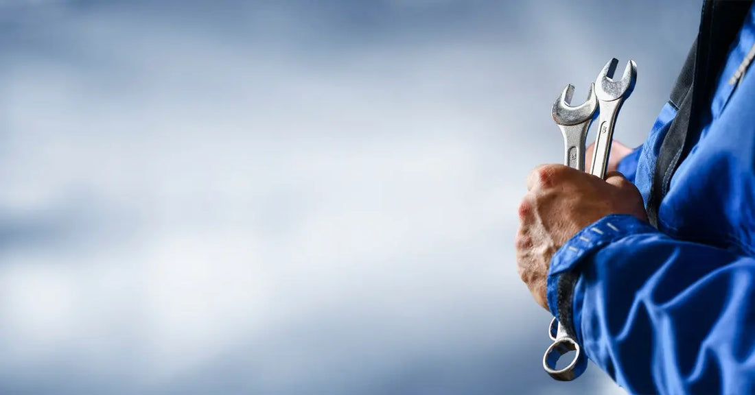 The side profile of an automotive mechanic in a blue jumpsuit holding a pair of silver wrenches up to his chest.