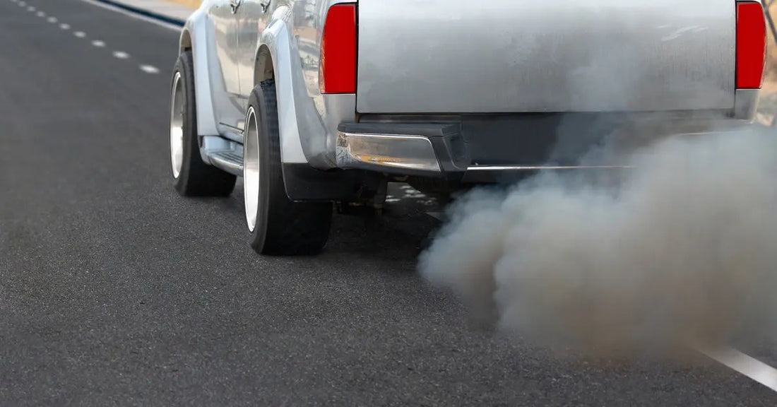 A pickup truck’s rear end on the road releases a thick gray cloud of exhaust smoke from the tailpipe.