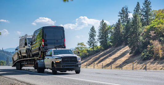 A pickup truck with a huge trailer attached drives on a highway, hauling two vans and one truck past pine-covered hills.