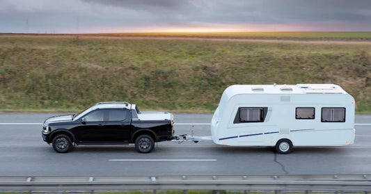 A black pickup truck towing a white travel trailer down a rural highway under a dark, stormy sky at dusk.