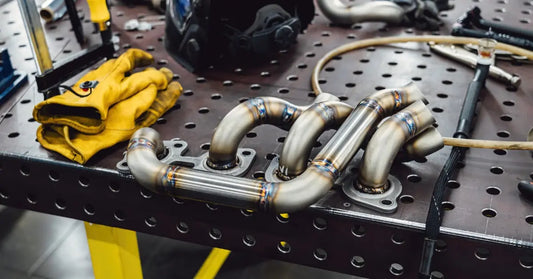 A welded stainless exhaust manifold rests on a metal workbench beside gloves, tools, and a black welding helmet.
