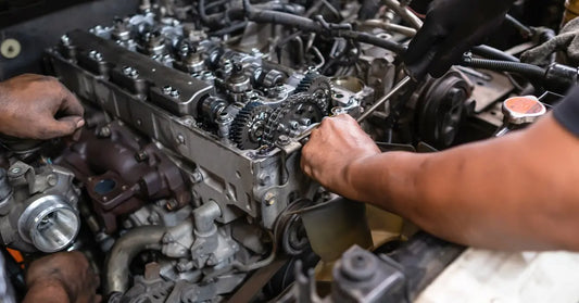 A mechanic using a screwdriver to remove nuts from the engine box of a diesel vehicle with its hood up.