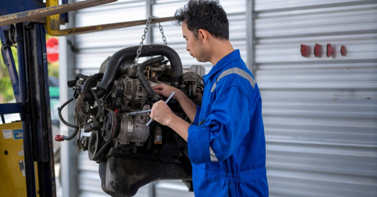 A mechanic in blue coveralls uses a wrench to repair a diesel engine hanging from a forklift chain in a garage bay.