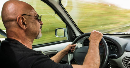 A bald man wearing sunglasses grips the steering wheel while driving his truck, the roadside blurred through the windows.