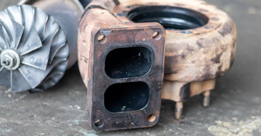 A rusted turbocharger housing with a rectangular twin-port flange sits beside a silver turbine wheel on a workbench.