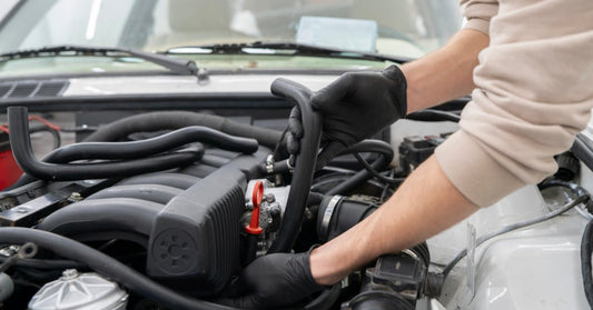A mechanic wearing black gloves positions a rubber hose inside a vehicle's engine bay near the air intake components.