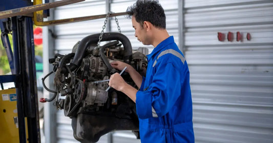 A mechanic in blue coveralls uses a wrench to repair a diesel engine hanging from a forklift chain in a garage bay.