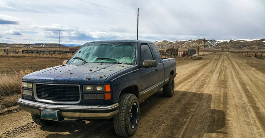 An older blue pickup truck is driving down a dirt road. The hood is covered in mud as it leaves a town behind it.